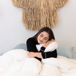 A woman enjoying a cozy night's sleep on a bed with pillows and a tiki, wrapped in the warmth of the Sienna Living Luxury Goose Feather Down Quilt.